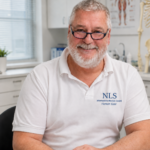Un hombre mayor sonriente, con gafas y barba blanca, en una consulta médica, vestido con un polo con el logotipo de NLS en el pecho.