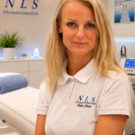 Portrait of a smiling blonde woman in a white polo shirt with the NLS logo, in a medical clinic.