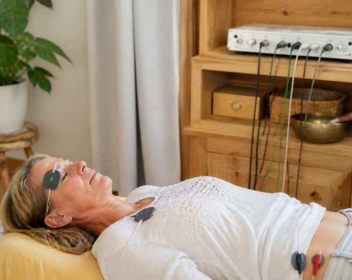 A woman lying on a massage-style table with EEG electrodes on her head and abdomen connected to a control device on a wooden shelf; a calm treatment session is underway.