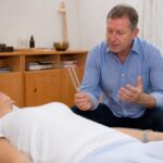 Frequency Therapy: Practical Guidance for Energy Therapists - 1790465372-1 - Herbert Eder - Frequency Researcher A male therapist is kneeling beside a reclining patient, holding a tuning fork and explaining the treatment in a clinic room.