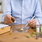 kombination-von-homoopathie-und-informationsmedizin-1789019563-1 - Herbert Eder - Frequenzforscher Person in blue shirt uses a dropper and a precision scale to measure small white pellets, with vials of pills on the table
