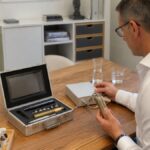 radionik-software-fur-heilpraktiker-richtig-anwenden-1788952288-1 - Herbert Eder - Frequenzforscher Man in a white shirt sits at a wooden table in a clinic, holding a handheld medical instrument next to an open silver case with tools inside the lid closed.