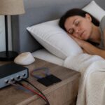 treating-sleep-disorders-naturally-with-frequencies-1790292981-1 - Herbert Eder - Frequency Researcher A woman sleeping in bed with an audio/monitoring device mounted on a nightstand and cables nearby.