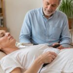 Frekvenční terapie pro vitalitu – aplikace v 6 krocích – 1790810920-1 – Herbert Eder – výzkumník v oblasti frekvencí Pacient středního věku ležící v posteli s elektrodami připevněnými na hrudníku, kterému lékař pomocí ručního přístroje podává elektrickou stimulaci.