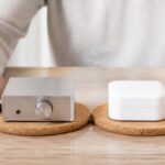 frequenztherapie-vs-placebo-mythos-aufgedeckt-2026-1790983628-1 - Herbert Eder - Frequenzforscher Silver portable amplifier with a knob and LED beside a white wireless earbud charging case on cork coasters.