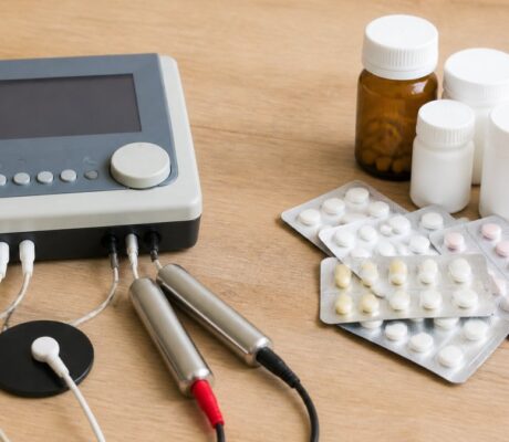 Frequency Therapy vs. Conventional Medicine for Pain-1791286668 - Herbert Eder - Frequency Researcher A medical device with electrodes and leads on a wooden table, next to blister packs of pills and pill bottles.