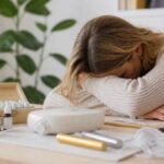 cos'è-la-medicina-informativa-nell'affaticamento-cronico-1792215632 - Herbert Eder - ricercatore nel campo delle frequenze Donna con la testa appoggiata sulle braccia, seduta a una scrivania su cui sono disposte delle bottiglie di oli essenziali e un diffusore.